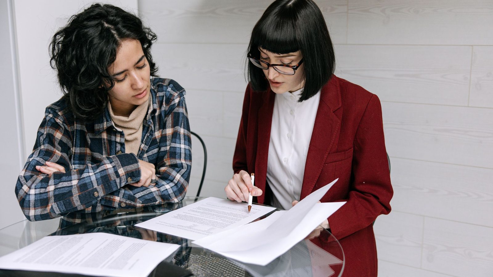 Two women collaborating at a glass table, reviewing business documents in a modern office setting