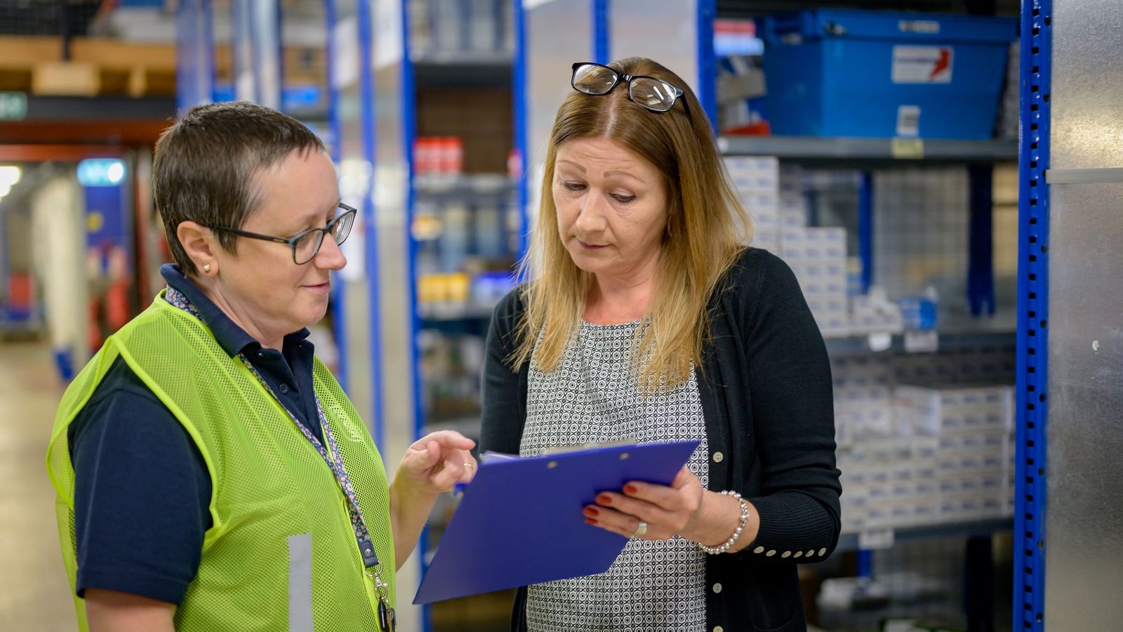 Two women in a warehouse reviewing an inventory checklist on a clipboard