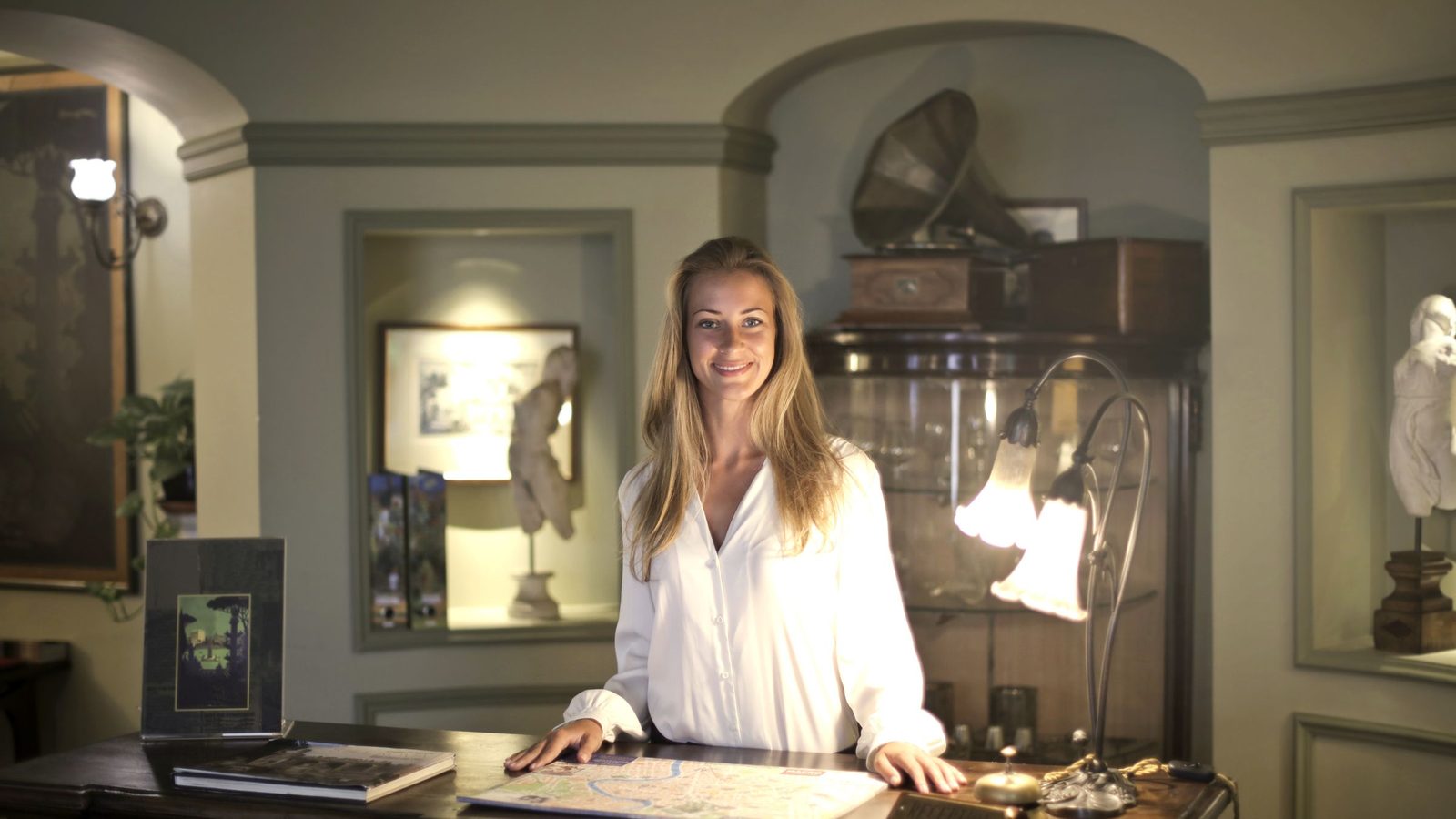 Smiling female receptionist wearing elegant clothes standing at reception desk in old fashioned interior of hotel and looking at camera