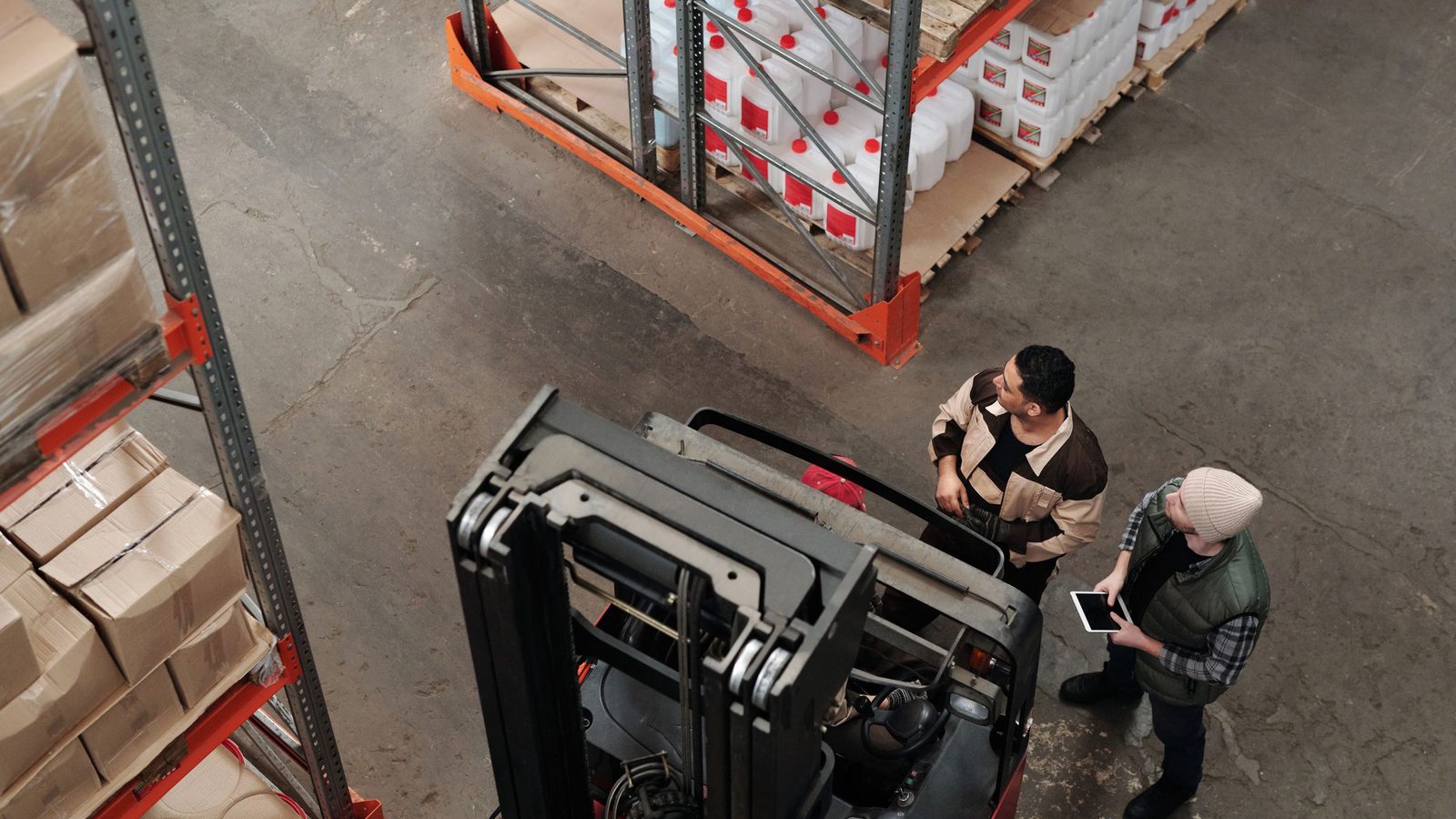 Two workers in a warehouse discussing logistics near a forklift captured from above