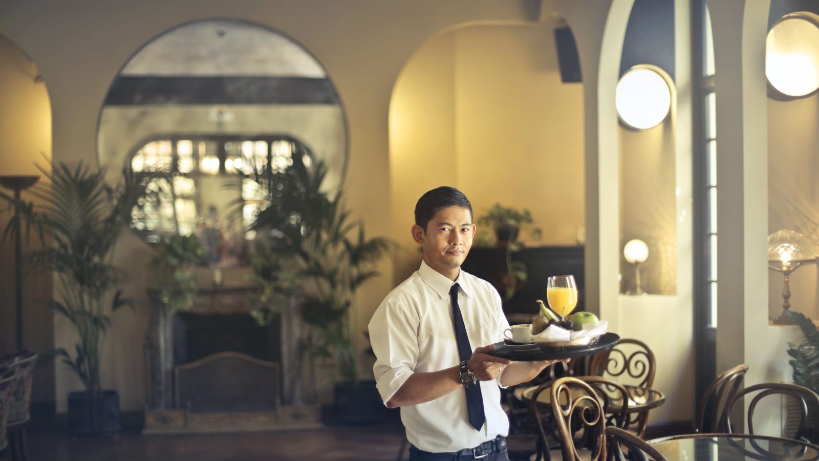 Confident young ethnic waiter in elegant clothes holding tray with food and drinks and looking at camera while serving tables in stylish restaurant